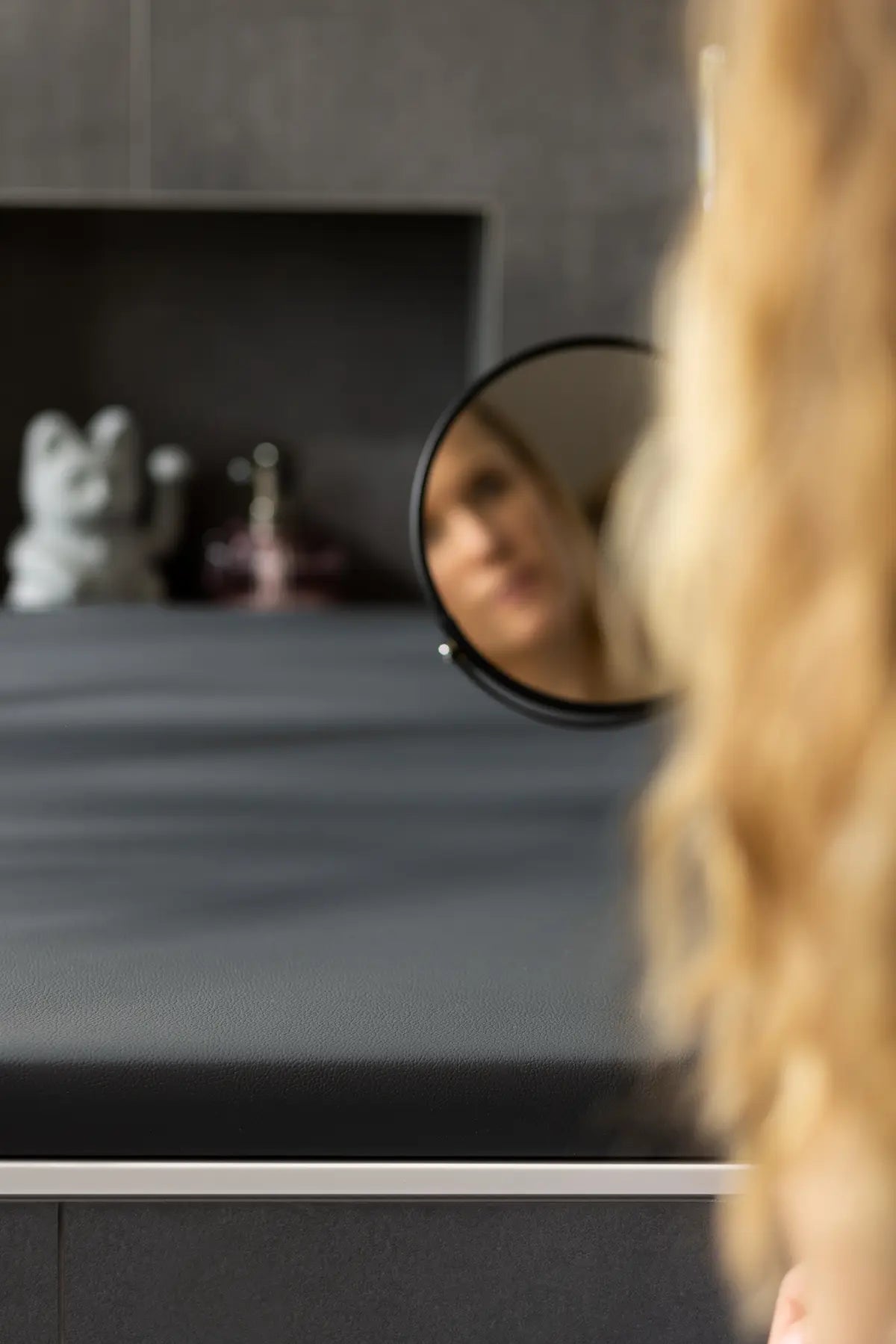 Das Spiegelbild einer Frau mit langen, blonden Haaren ist in einem runden Handspiegel zu sehen, der über einer anthrazitfarbenen Badewannenabdeckung bzw. Abdeckung Badewanne liegt. Im Hintergrund befinden sich eine dekorative Figur und eine stilvolle Parfümflasche, die die elegante Ästhetik des Raums betonen.