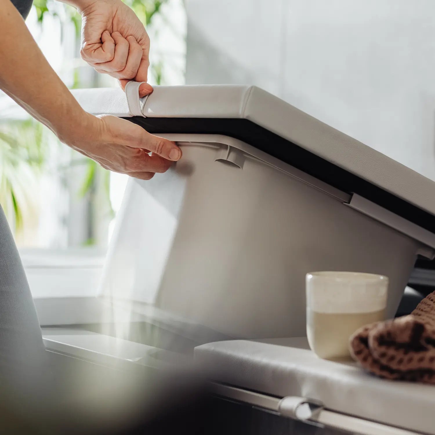 Eine Person hebt den Deckel der hejBox in hellgrauer Farbe an, um den versteckten Stauraum in der Badewanne zu nutzen. Die hejBox, eine durchdachte Aufbewahrungslösung, fügt sich perfekt in das minimalistische Badezimmerdesign ein. Neben der Badewanne steht eine Tasse, und ein strukturiertes Textil setzt einen stilvollen Akzent, der Funktionalität mit modernem Design verbindet. Bestelle die Matti Badewannenabdeckung bzw. Abdeckung Badewanne.