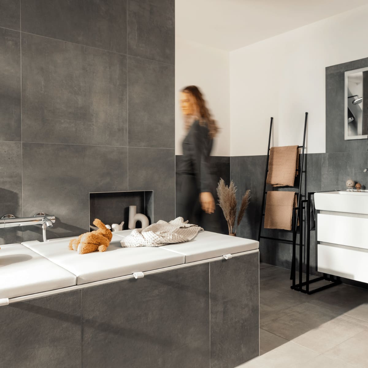 Ein gut beleuchtetes Badezimmer mit einer Badewannenabdeckung in ashgrey, auf der ein Kuscheltier und eine gefaltete Decke liegen. Ein Mann in blauer Nachtwäsche steht vor einem großen Spiegel am Waschtisch und putzt sich die Zähne, während eine Frau im Hintergrund an der Badewanne vorbeigeht. Die Badezimmereinrichtung umfasst einen modernen Waschtisch, Handtuchhalter und dezente Dekorationen, die für eine ordentliche und komfortable Atmosphäre sorgen.