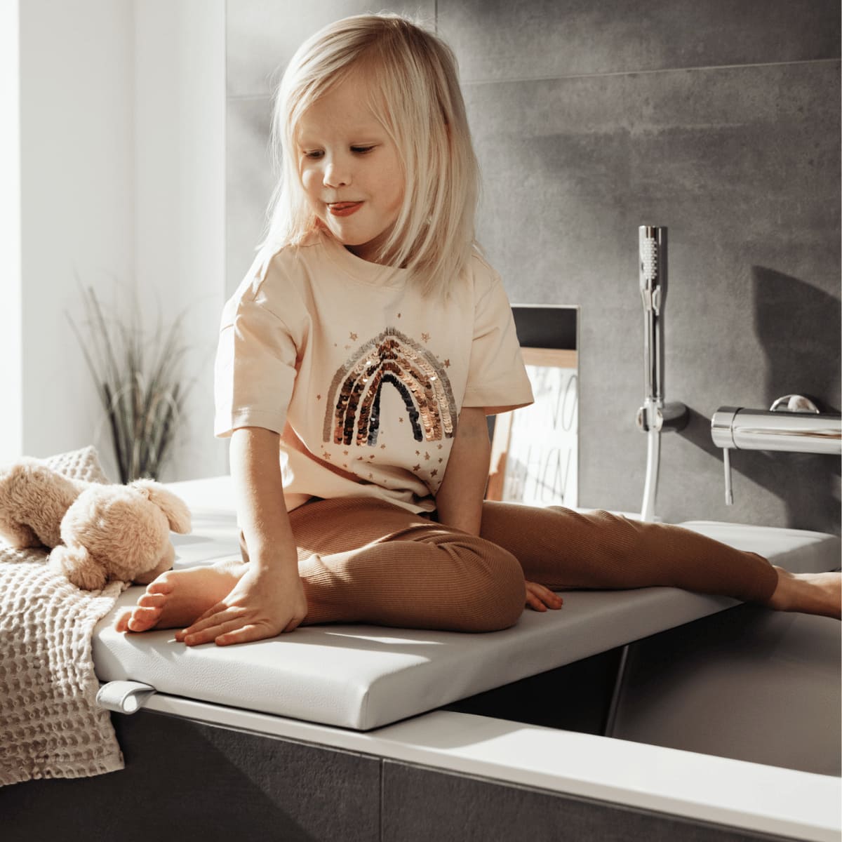 Ein kleines Kind sitzt fröhlich auf einer Badewannenabdeckung bzw. Abdeckung Badewanne in hellem Grau und spielt mit einem Kuscheltier. Das Kind trägt ein T-Shirt mit einem funkelnden Regenbogenmotiv und ist umgeben von weichen Textilien und einer sanft beleuchteten Badezimmerumgebung. Die Szene strahlt Wärme und Geborgenheit aus und zeigt, wie sicher und bequem die Abdeckung im Alltag genutzt werden kann.