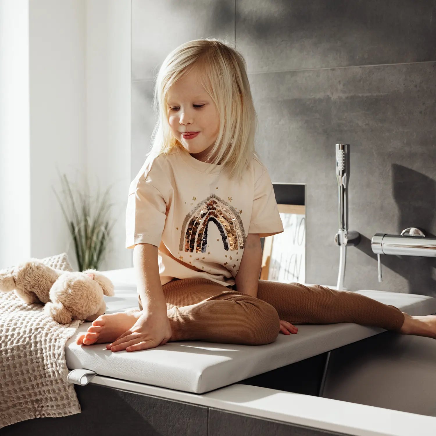 Ein kleines Kind sitzt fröhlich auf einer Badewannenabdeckung bzw. Abdeckung Badewanne in hellem Grau und spielt mit einem Kuscheltier. Das Kind trägt ein T-Shirt mit einem funkelnden Regenbogenmotiv und ist umgeben von weichen Textilien und einer sanft beleuchteten Badezimmerumgebung. Die Szene strahlt Wärme und Geborgenheit aus und zeigt, wie sicher und bequem die Abdeckung im Alltag genutzt werden kann.