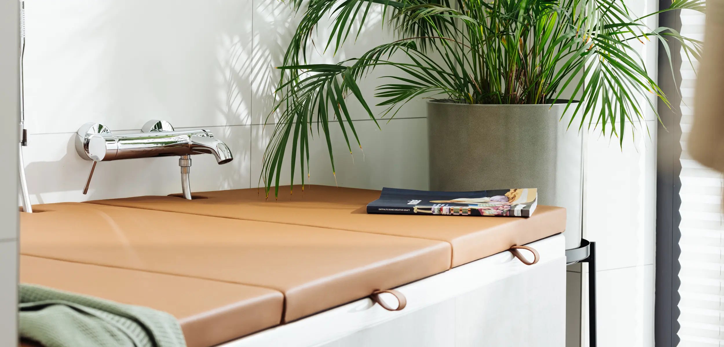 Ein modernes Badezimmer mit einer Badewannenabdeckung bzw. Abdeckung Badewanne in warmem Cognac-Braun, die als zusätzliche Ablagefläche dient. Auf der Abdeckung liegen ein aufgeschlagenes Magazin und ein Handtuch, während eine große Zimmerpflanze dem Raum Frische und Lebendigkeit verleiht. Die durchdachte Farbwahl und das minimalistische Design der Abdeckung fügen sich harmonisch in die Gesamtgestaltung des Badezimmers ein.