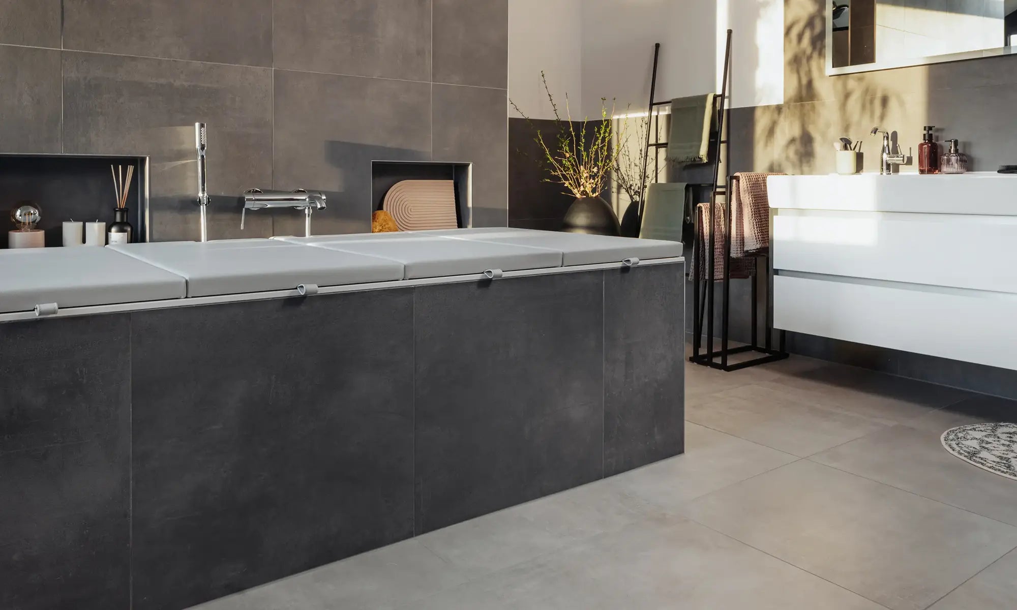 Eine elegante Matti Badewannenabdeckung bzw. Abdeckung Badewanne in ashgrey verleiht diesem modernen Badezimmer ein stilvolles, minimalistisches Aussehen. Das sanfte Grau harmoniert perfekt mit den dunklen Fliesen und der modernen Einrichtung, während die goldene Abendsonne den Raum in ein warmes Licht taucht