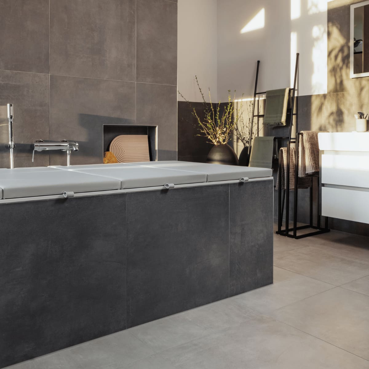 Eine elegante Matti Badewannenabdeckung bzw. Abdeckung Badewanne in ashgrey verleiht diesem modernen Badezimmer ein stilvolles, minimalistisches Aussehen. Das sanfte Grau harmoniert perfekt mit den dunklen Fliesen und der modernen Einrichtung, während die goldene Abendsonne den Raum in ein warmes Licht taucht