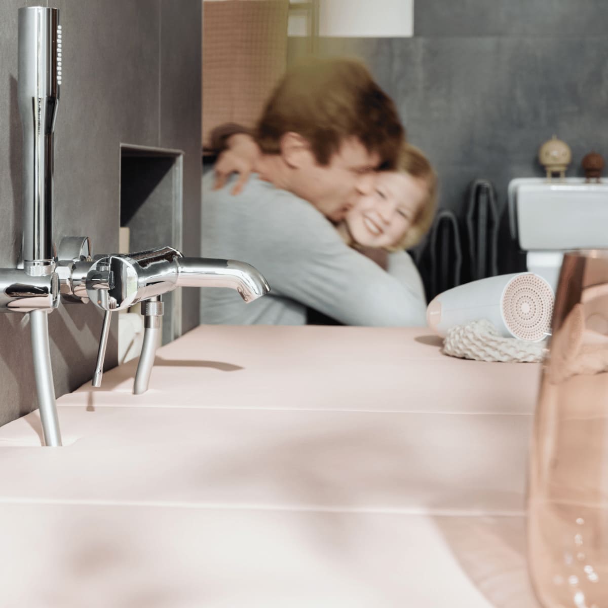 Im Vordergrund eines modernen Badezimmers mit einer zartrosa Badewannenabdeckung bzw. Abdeckung Badewanne sind glänzende Chromarmaturen zu sehen. Im Hintergrund umarmt ein Vater liebevoll seine Tochter, was dem Raum eine warme, familiäre Atmosphäre verleiht. Neben der Badewanne liegen ein zusammengerolltes Textil und ein Fön, die das moderne und funktionale Design des Badezimmers ergänzen.