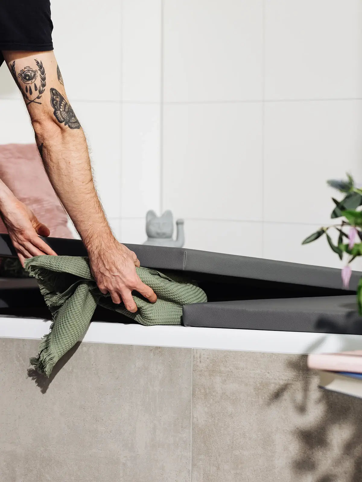 Ein Mann mit tätowiertem Arm hebt eine matte, dunkelgraue Badewannenabdeckung bzw. Abdeckung Badewanne an, um ein grünes Tuch im Stauraum darunter zu verstauen. Neben der Badewanne steht ein Tisch mit frischen Blumen, die dem Badezimmer einen Hauch von Farbe und Natur verleihen. Die Kombination aus praktischer Funktionalität und stilvollem Design unterstreicht die durchdachte Einrichtung des Raums.