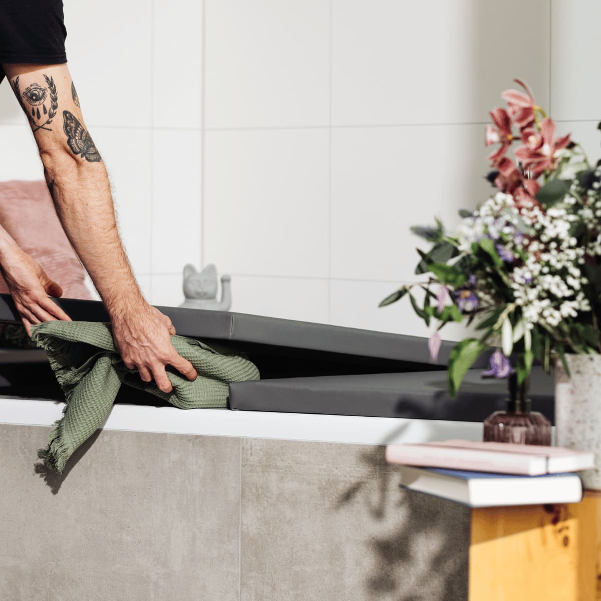 Eine Person mit tätowiertem Arm legt ein grün gefaltetes Tuch in eine geöffnete Badewannenabdeckung bzw. Abdeckung Badewanne in der Farbe Slate Grey. Im Vordergrund steht eine Vase mit bunten Blumen, daneben liegen Bücher auf einem Holztisch. Im Hintergrund ist eine graue Wand mit einer Dekofigur zu sehen. Die Szene unterstreicht den praktischen Stauraum, den die Abdeckung bietet, und verbindet Ästhetik mit Funktionalität.