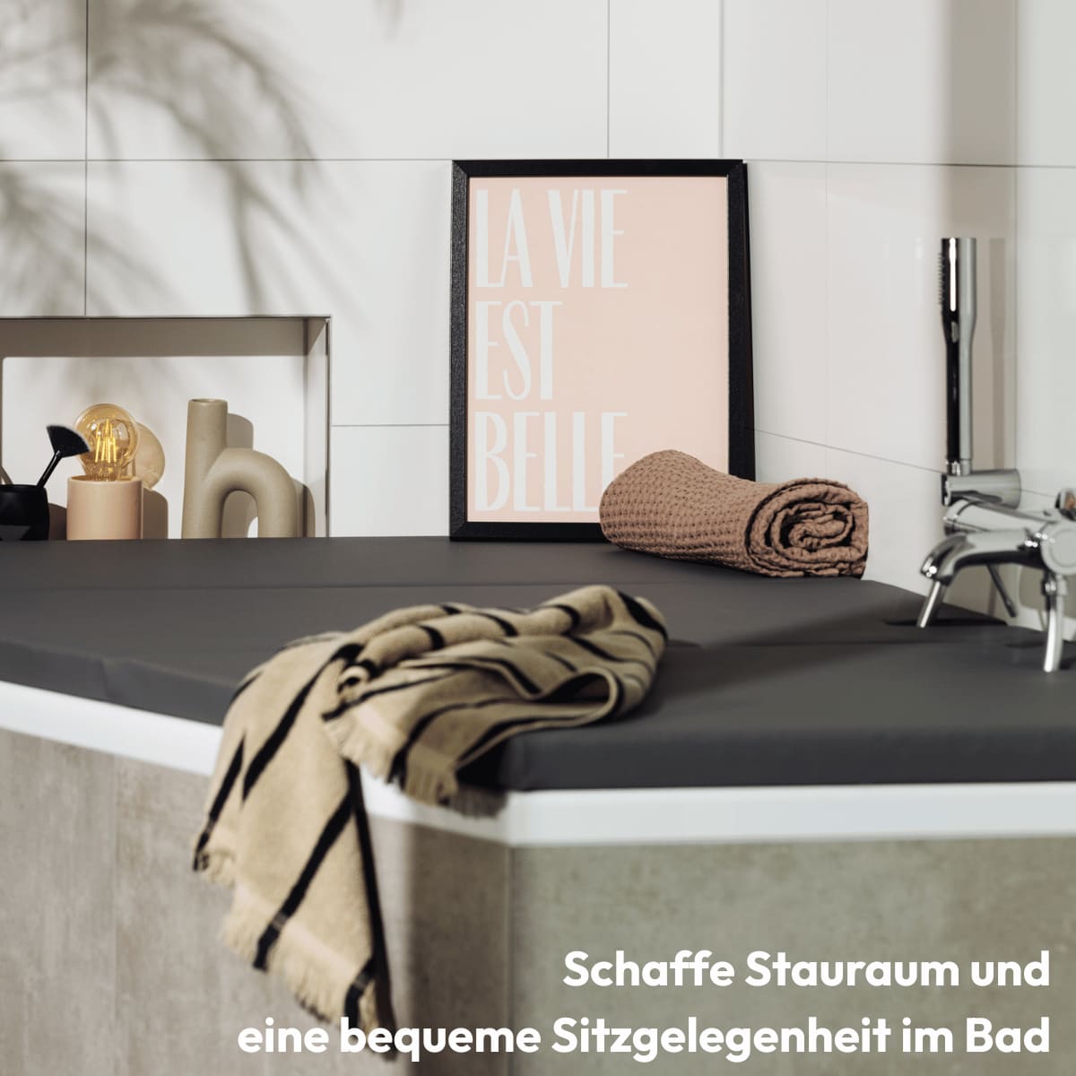 Eine moderne Badezimmerabdeckung in der Farbe slate grey, die stilvoll über einer Badewanne liegt. Auf der Abdeckung befinden sich ein gerolltes Handtuch, ein brauner Seifenspender, eine aufgeschlagene Zeitschrift und ein kunstvoll gerahmtes Bild mit der Aufschrift "La Vie Est Belle". Die Szene wird durch sanfte Lichtspiele und eine ruhige Atmosphäre unterstrichen, was den Raum zu einem harmonischen Rückzugsort macht. Bestelle deine Matti Badewannenabdeckung bzw. Abdeckung Badewanne.