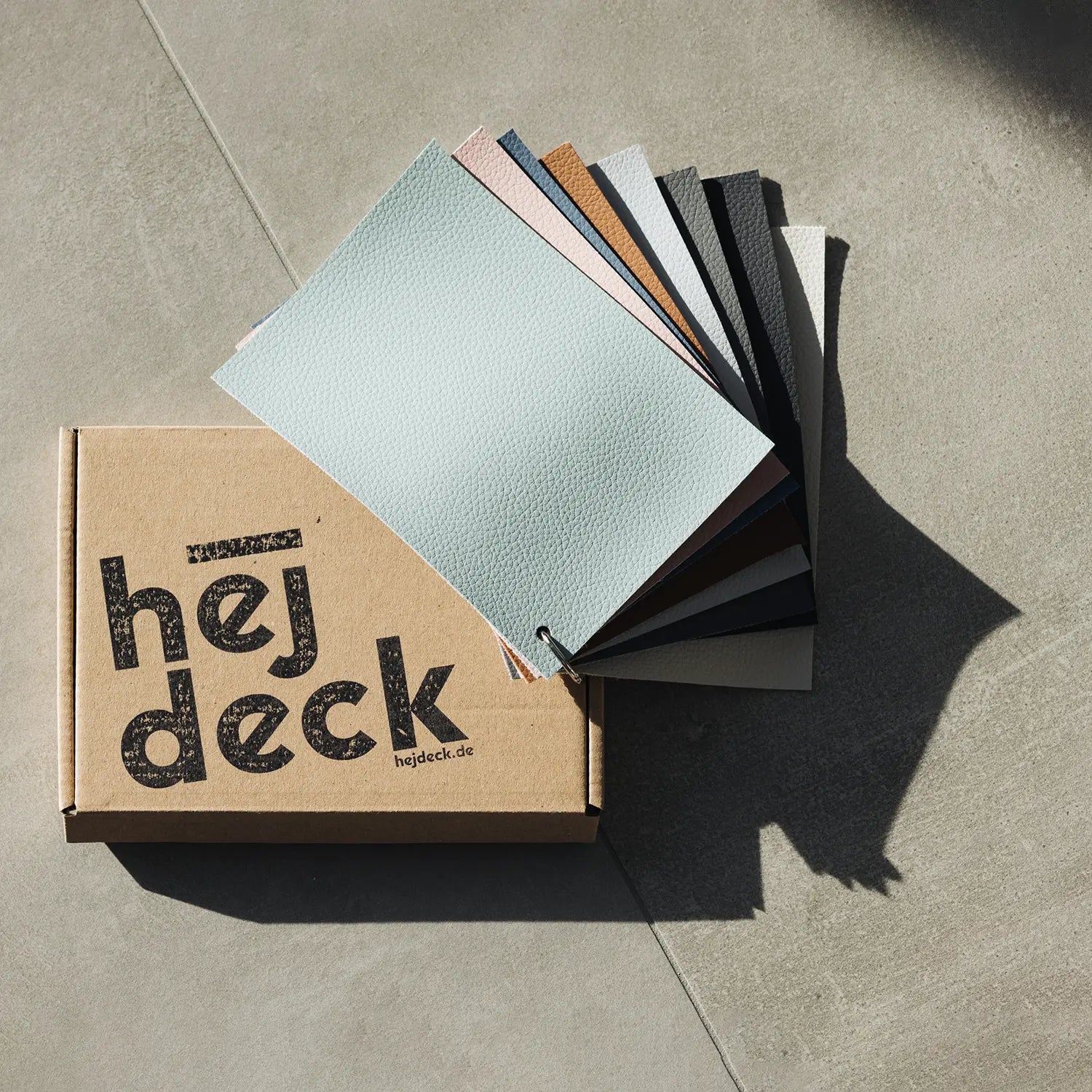 Die kostenfreie hejdeck-Musterbox mit allen acht angebotenen Farben liegt geöffnet auf einem hellen Betonboden. Der enthaltene Musterfächer zeigt die verschiedenen Farbtöne und Texturen, die für Badewannenabdeckungen verfügbar sind. Die schlichte Kartonschachtel trägt das hejdeck-Logo, was die hochwertige Qualität und die Auswahlmöglichkeiten der hejdeck-Produkte unterstreicht.