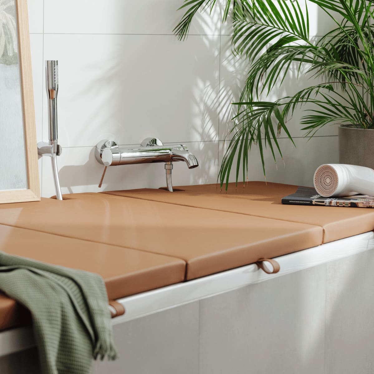 Stilvolle Badewannenabdeckung bzw. Abdeckung Badewanne in cognacbraun, die als Sitzfläche und Ablagefläche dient. Die Abdeckung ist maßgefertigt und fügt sich harmonisch in ein modernes Badezimmer ein, umgeben von einer grünen Pflanze und dekorativen Details wie einem Handtuch und einem Fön.