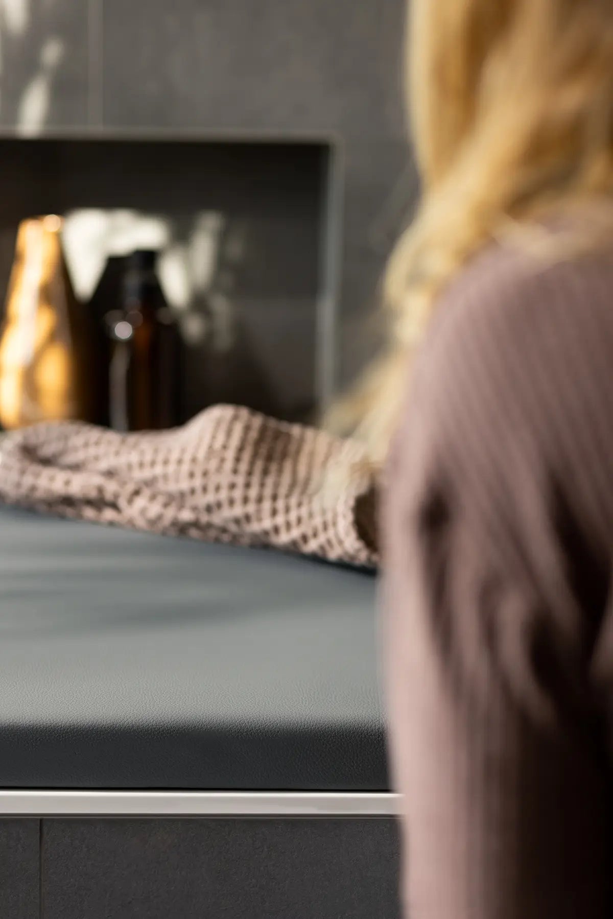Eine Person in einem beige-pinken Pullover sitzt am Rand einer präzise zugeschnittenen Badewannenabdeckung Holz bzw. Abdeckung Badewanne in slate grey. Ein weiches, gestricktes Handtuch liegt auf der Oberfläche, während im Hintergrund stilvolle, minimalistische Badeaccessoires in warmen Gold- und Brauntönen zu sehen sind.