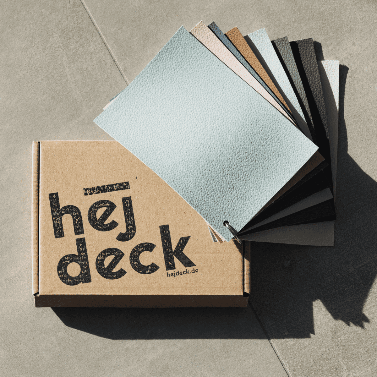 Die kostenfreie hejdeck-Musterbox mit allen acht angebotenen Farben liegt geöffnet auf einem hellen Betonboden. Der enthaltene Musterfächer zeigt die verschiedenen Farbtöne und Texturen, die für Badewannenabdeckungen verfügbar sind. Die schlichte Kartonschachtel trägt das hejdeck-Logo, was die hochwertige Qualität und die Auswahlmöglichkeiten der hejdeck-Produkte unterstreicht.