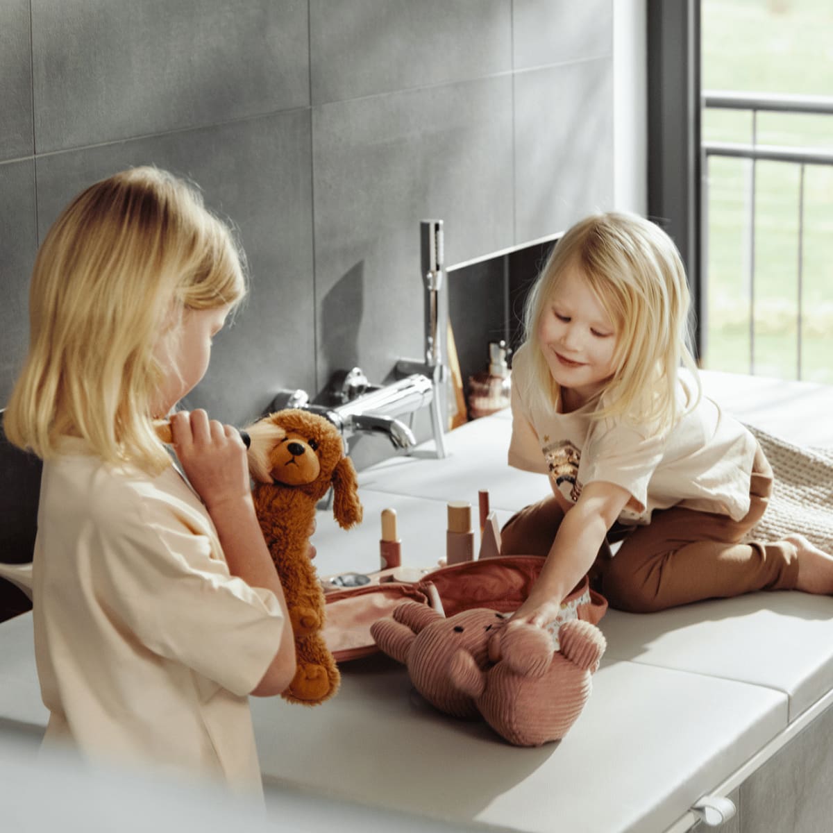 Zwei Kinder spielen auf einer cremeweißen Matti-Badewannenabdeckung von hejdeck mit Kuscheltieren und Spielzeug. Die Abdeckung verwandelt die Badewanne in eine sichere Spielfläche und zeigt, wie vielseitig nutzbar der zusätzliche Stauraum und die robuste Oberfläche im Alltag sind.