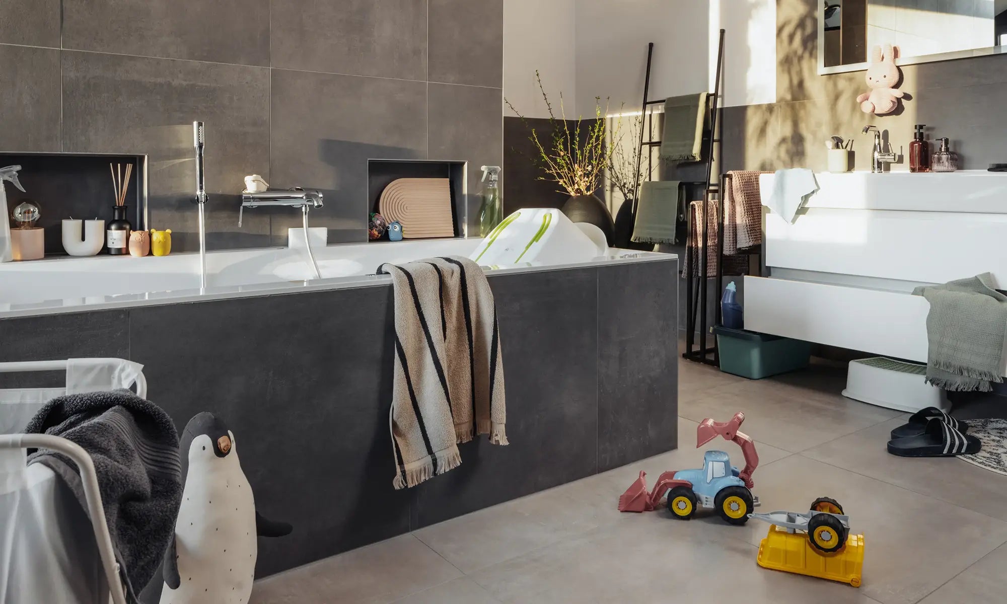 Ein unordentliches Badezimmer mit einer eingebauten Badewanne, umgeben von verstreutem Kinderspielzeug und Badeutensilien. Handtücher und Kleidung sind wahllos auf dem Boden und über den Rand der Badewanne verteilt, während es an Stauraummöglichkeiten fehlt. Die fehlende Aufbewahrungslösung lässt wenig Platz für schicke Dekorationen und Ordnung